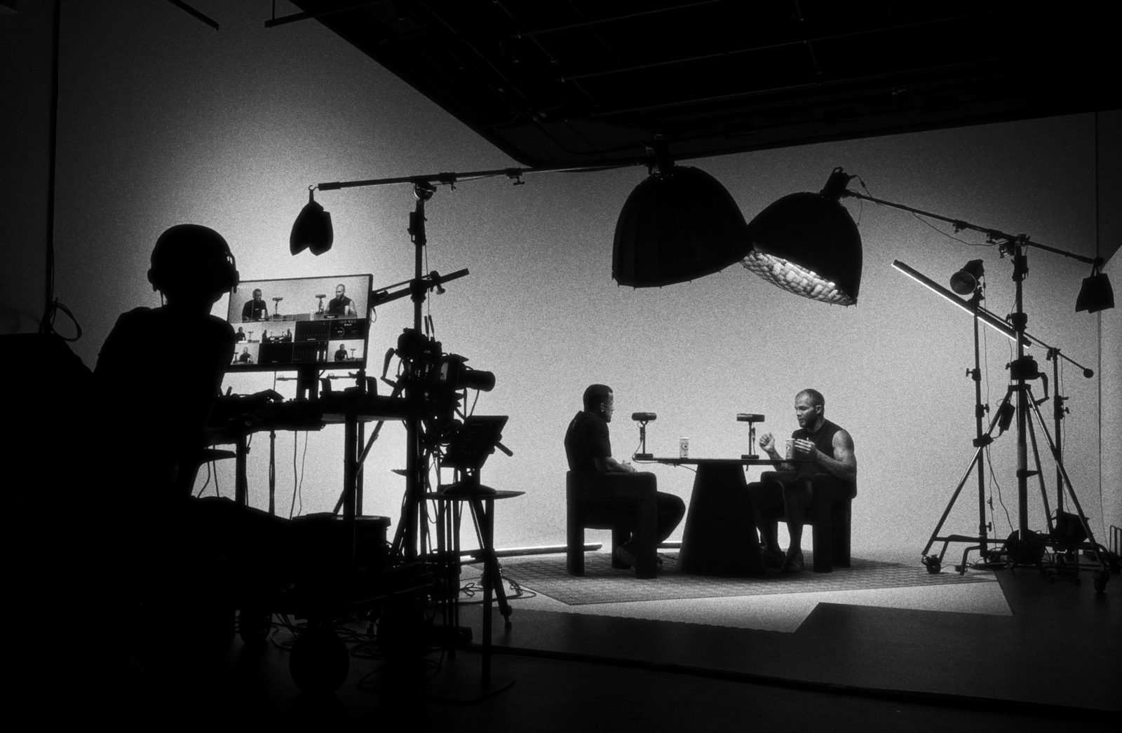 A two-camera podcast set in a London studio: two guests at a table under softboxes, director at the multiview monitor.